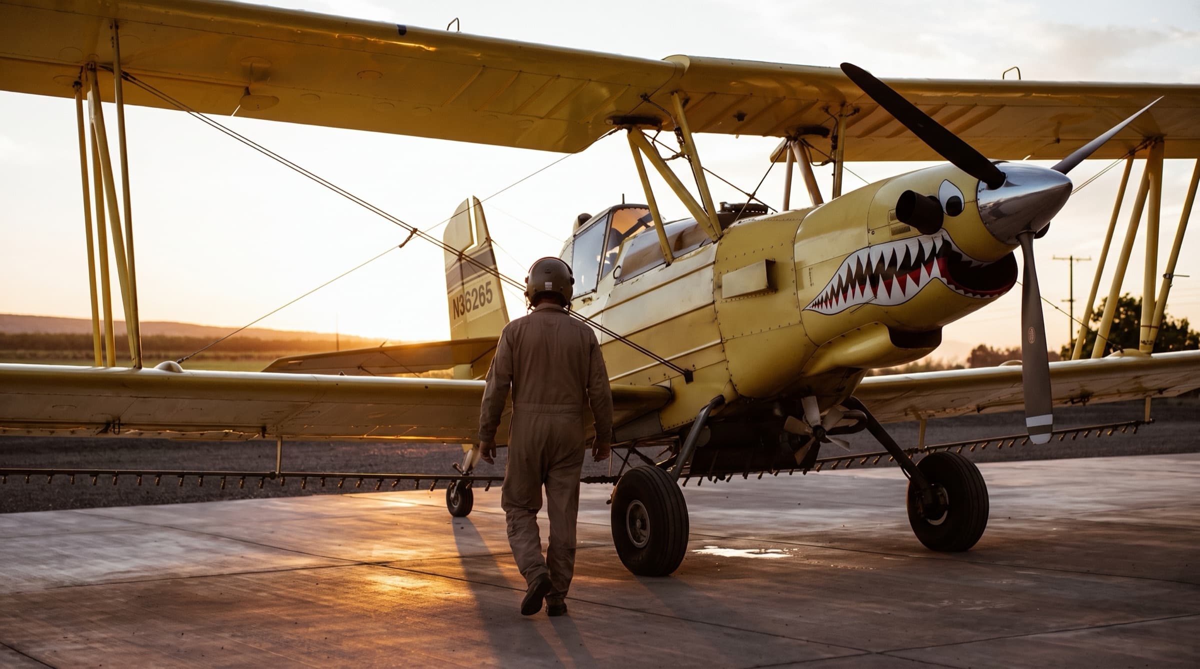An ag pilot in a flight helmet and coveralls walks to a yellow Ag Cat biplane on the ramp at sunrise.